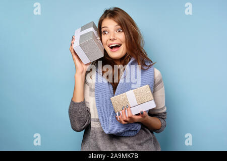 A brunette in a sweater and scarf stands on a blue background with gift boxes, putting one to ear with a joyful expression on face Stock Photo