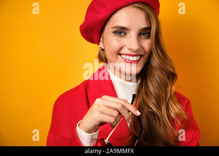 Portrait of a smiling blonde with red lipstick, in a white blouse, red coat and beret with glasses in his hand, focus on the face Stock Photo