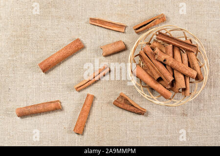 Heap of cinnamon bark sticks in wicker bowl, some scattered on linen tablecloth, view from above Stock Photo