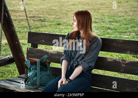 Red-haired girl sits with a backpack on a wooden bench swing and looks away. Stock Photo