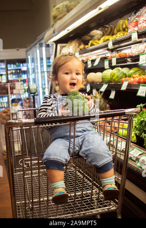 A smiling baby munches broccoli in the produce isle of a grocery