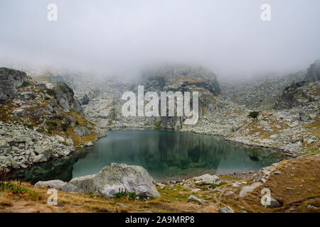 View of a beautiful high mountain lake with calm water surrounded by foggy rocky shore. The Scary Lake, Rila National Park, Bulgaria.