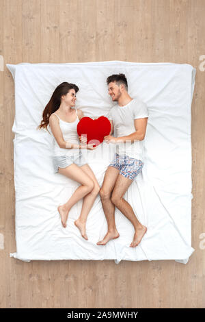 Bedtime. Young couple lying on bed top view holding heart shape pillow looking at each other smiling cheerful Stock Photo