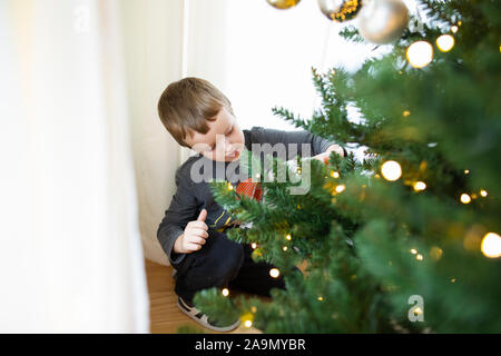 Young boy kneels to put ornament on lit Christmas tree branch Stock Photo