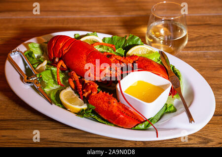 Cooked lobster on a white ceramic platter, romaine lettuce, lemon slices, melted butter, and white wine on the side in a stemless wine glass Stock Photo