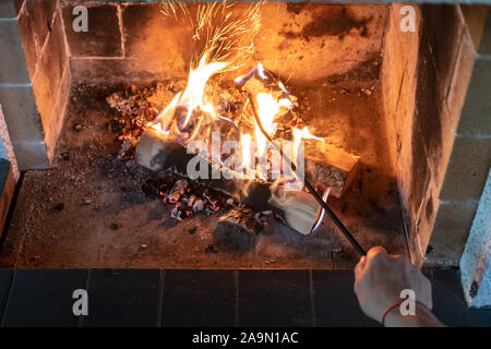 Female hand straightens and overturns burning firewood with an iron poker in an open fireplace and from the coal fly beautiful spark. Warmth and Stock Photo