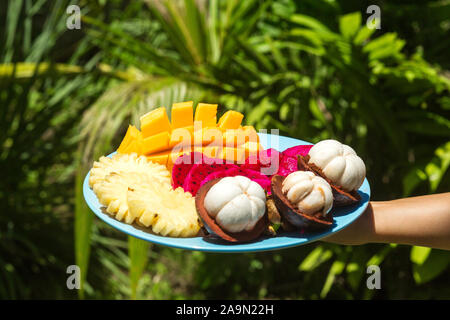 A girl in the green jungle is holding a plate with freshly cut and peeled tropical fruits. Stock Photo