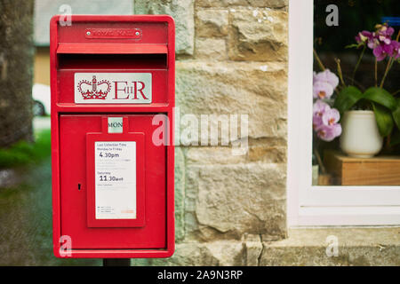 Red British post box with 'ER' symbol in front of a stone building with window in it displaying flowers Stock Photo