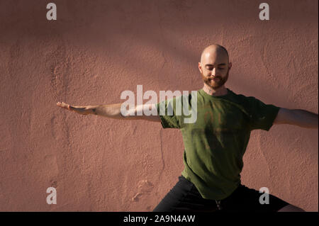 Young man in his thirties with a beard wearing a green tee shirt practicing yoga by a salmon pink colored adobe wall. Stock Photo