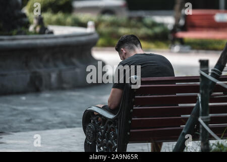 A young white man sits alone on a bench looking down feeling sad Stock Photo
