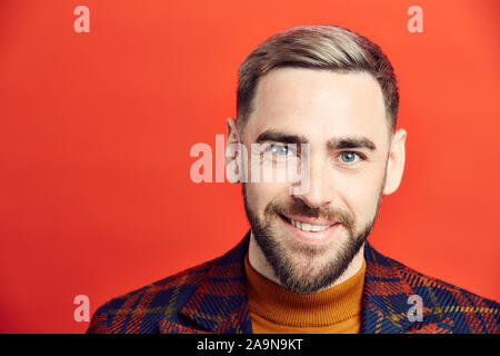 Head and shoulders portrait of smiling bearded man looking at camera while posing against red background, copy space Stock Photo