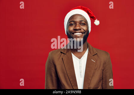 Waist up portrait of happy African-American man wearing Santa hat smiling at camera while standing against red background, copy space Stock Photo