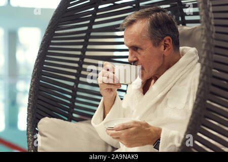 Side view portrait of wealthy senior man relaxing in lounge chair at luxury SPA resort and drinking coffee Stock Photo