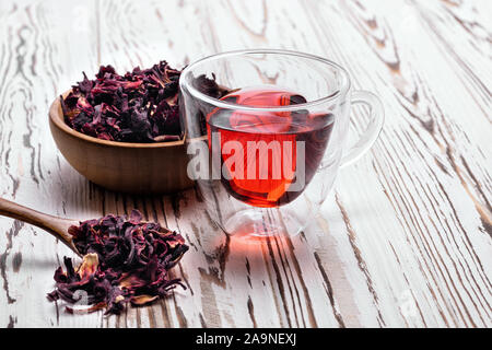 red hibiscus tea in glass cup on white wooden table with dry rose petals in spoon and bowl Stock Photo