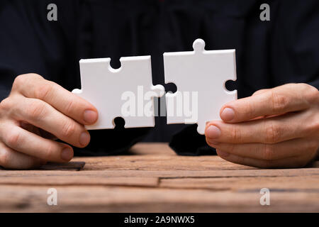 Person's Hand Joining White Jigsaw Puzzle Pieces On Wooden Surface Stock Photo