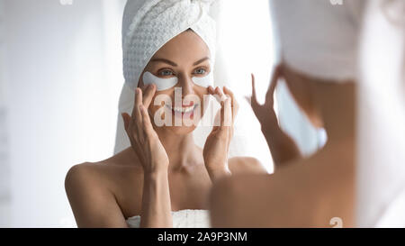 Happy woman towel on head apply patches looking in mirror Stock Photo