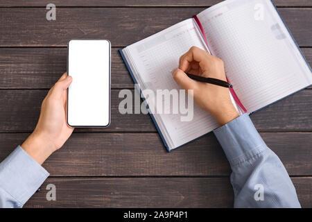 Male hand holding blank smartphone and writing in notebook Stock Photo