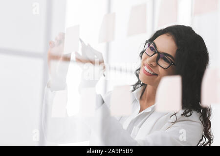 Mexican woman writing ideas on sticky notes over glass wall Stock Photo