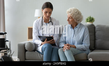 Young female doctor consulting disabled old patient writing in clipboard Stock Photo