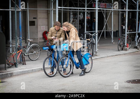 A well dressed couple, possibly tourists, sit on their Citi Bikes while looking at a cell phone. On Union Square West in lower Manhattan, New York Cit. Stock Photo
