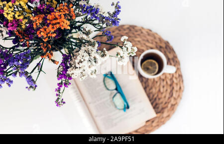 bouquet of flowers, open book and glasses, lemon tea in a white ceramic cup on a wicker stand on the table, top view Stock Photo