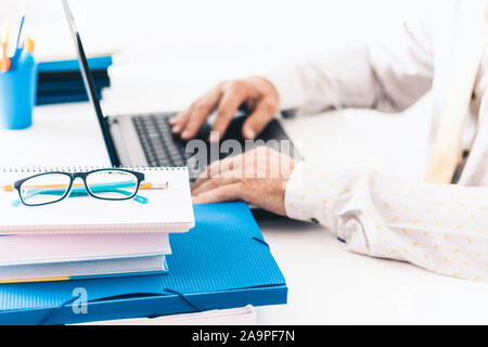 Close-up of hands middle-aged man in white shirt typing on keyboard laptop computer, holder with pencils and pens, stack of books, notebooks, smartpho Stock Photo