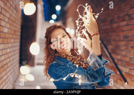 Happy young woman playing with fairy lights outdoors in city, toothy smile