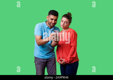 Portrait of curious attractive young couple in casual wear standing, reading message together on mobile phone, looking concentrated and interested. is Stock Photo