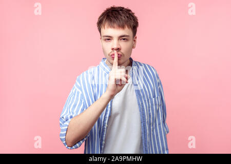 Be quiet! Portrait of young brown-haired man with small beard and mustache in casual striped shirt making silence gesture, saying hush, keep silence. Stock Photo