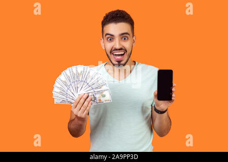 Online payment. Portrait of extremely happy man with beard in white t-shirt holding money and cellphone, looking with amazement, pleasantly surprised.