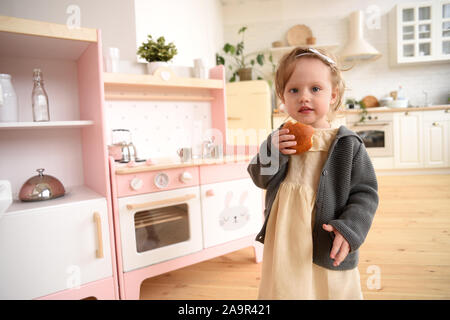 Little child with sweets concept. Cute toddler girl eating bun near play kitchen in light empty room copy space Stock Photo