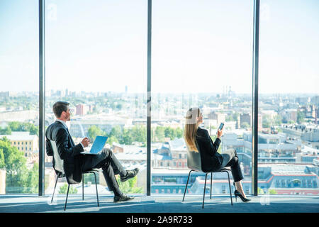 Business man with laptop and business woman with smartphone sitting by the window in panoramic office Stock Photo