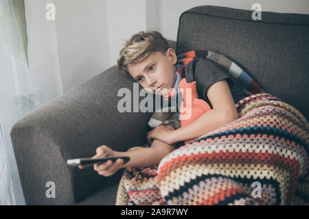 Sick boy watching tv with woolen blanket and hot water bottle. Sad teen with the flu rests at home in a cold winter day. Child with seasonal infection Stock Photo