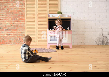 Family and children concept. Toddler boy playing on floor and cute little girl eating sweets at play kitchen in big empty room copy space Stock Photo