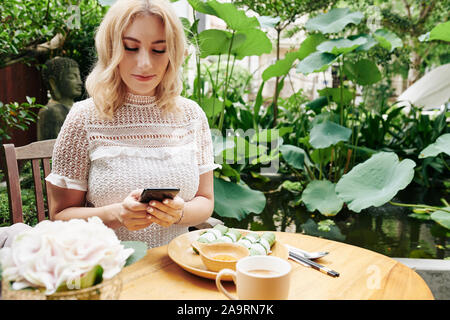 Positive young blond woman eating tasty lunch and texting friends when sitting at table outdoors Stock Photo