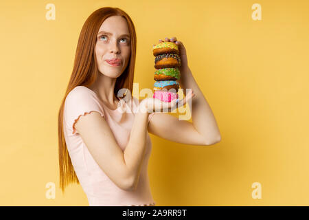 Shot of happy caucasian woman with red hair, closed eyes, licking her lips with tongue, holding in hands delicious colorful donuts isolated over yello Stock Photo
