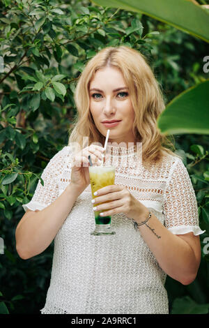 Portrait of young beautiful woman in white lace dress drinking refreshing fruit cocktail and looking at camera Stock Photo