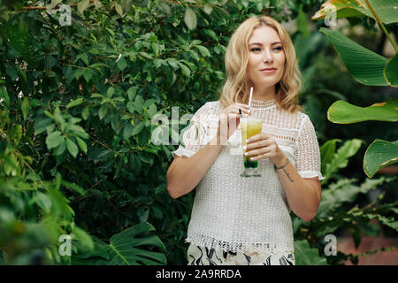 Young attractive Caucasian woman enjoying delicious fruit cocktail when standing in garden among green trees Stock Photo