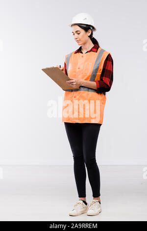 Pretty female builder in hardhat, flannel, leggins and working vest making notes Stock Photo