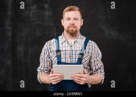 Young successful technician in checkered shirt and blue overalls using tablet Stock Photo