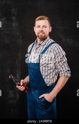 Happy young bearded mechanic in coveralls and shirt using handtool while working Stock Photo