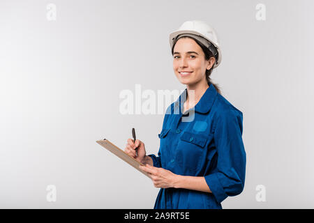 Happy young female builder in blue workwear and hardhat making notes in document Stock Photo
