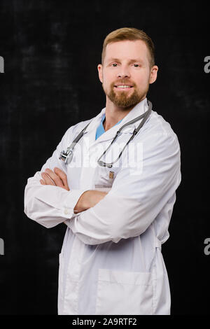 Happy young bearded doctor in whitecoat crossing arms on chest in isolation Stock Photo