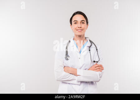 Pretty young smiling clinician in whitecoat crossing arms on chest in isolation Stock Photo