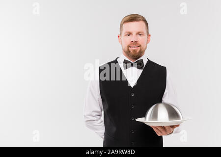 Happy young waiter in waistcoat and bowtie holding cloche with meal Stock Photo