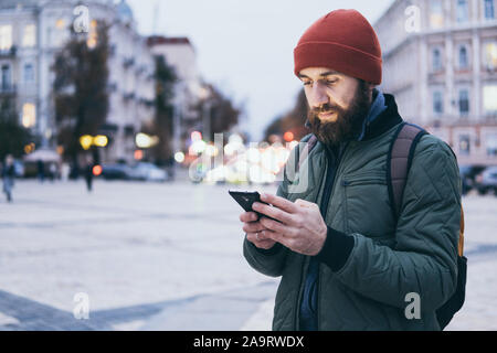 Young bearded Caucasian man looking on mobile phone screen while walking on the street Stock Photo