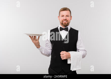 Young elegant waiter in black waistcoat and bowtie holding white towel and plate Stock Photo