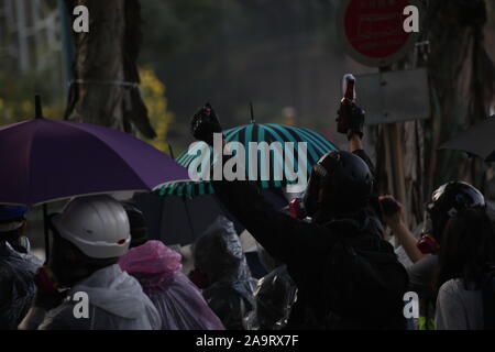 Hong Kong, China. 17th Nov 2019. A protestor clad in black raises his hands jubilantly while heckling the police at Polytechnic University, a home base for many of the student protestors in Hong Kong for the past week. He holds a petrol bomb in one hand, a weapon that has been used with increasing frequency by protestors since the movement first started in July 2019. Umbrellas can also be seen in the background, which have been a symbolic and strategic tool of both the 2014 Umbrella Revolution and 2019 Hong Kong Protests. Credit: Katherine Cheng/Alamy Live News. Stock Photo