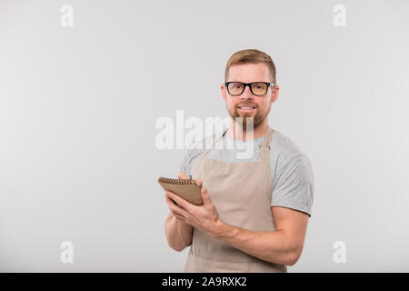 Happy young bearded waiter writing down order of client in notepad Stock Photo