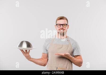 Happy young bearded waiter in apron holding cloche with cooked food Stock Photo
